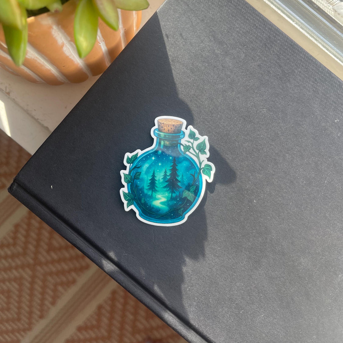 Sticker of a blue potion bottle on a black surface with a plant in the background