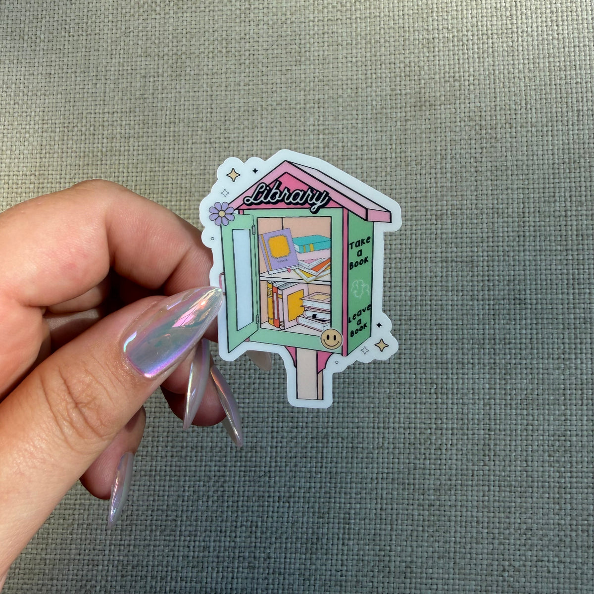 A hand with iridescent nail polish holds a die-cut sticker shaped like a pastel library bookshelf against a textured gray fabric background.