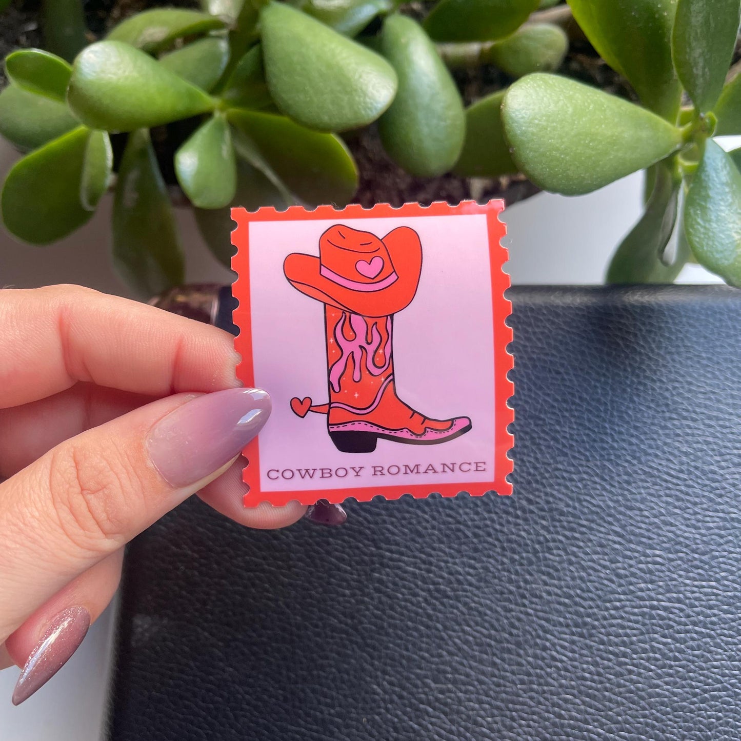 Hand holding a card with a cowboy boot and hat illustration labeled 'Cowboy Romance' in front of green plants.