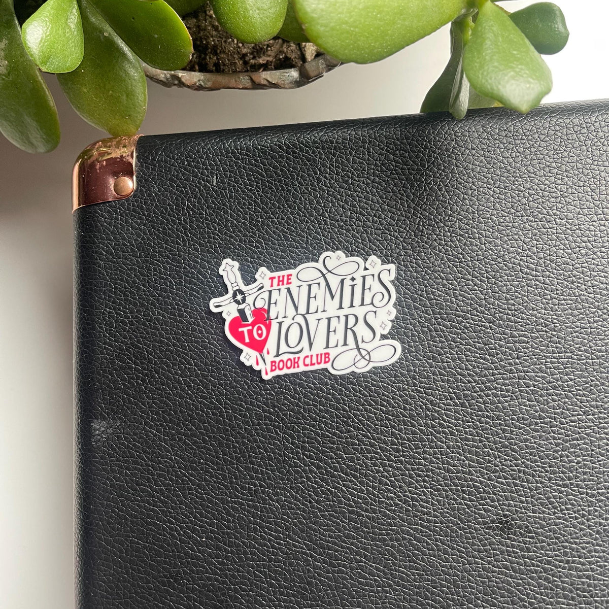 Black leather notebook with a sticker on it, surrounded by green plants.