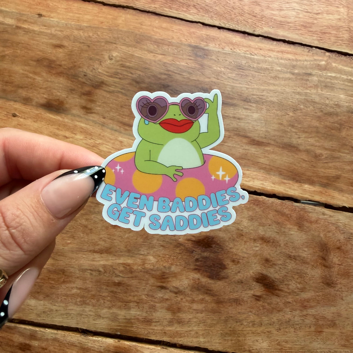 A hand with manicured nails holds a die-cut sticker featuring a green cartoon frog wearing heart-shaped sunglasses and a pink float with the text 'EVEN BADDIES. GET SADDIES.