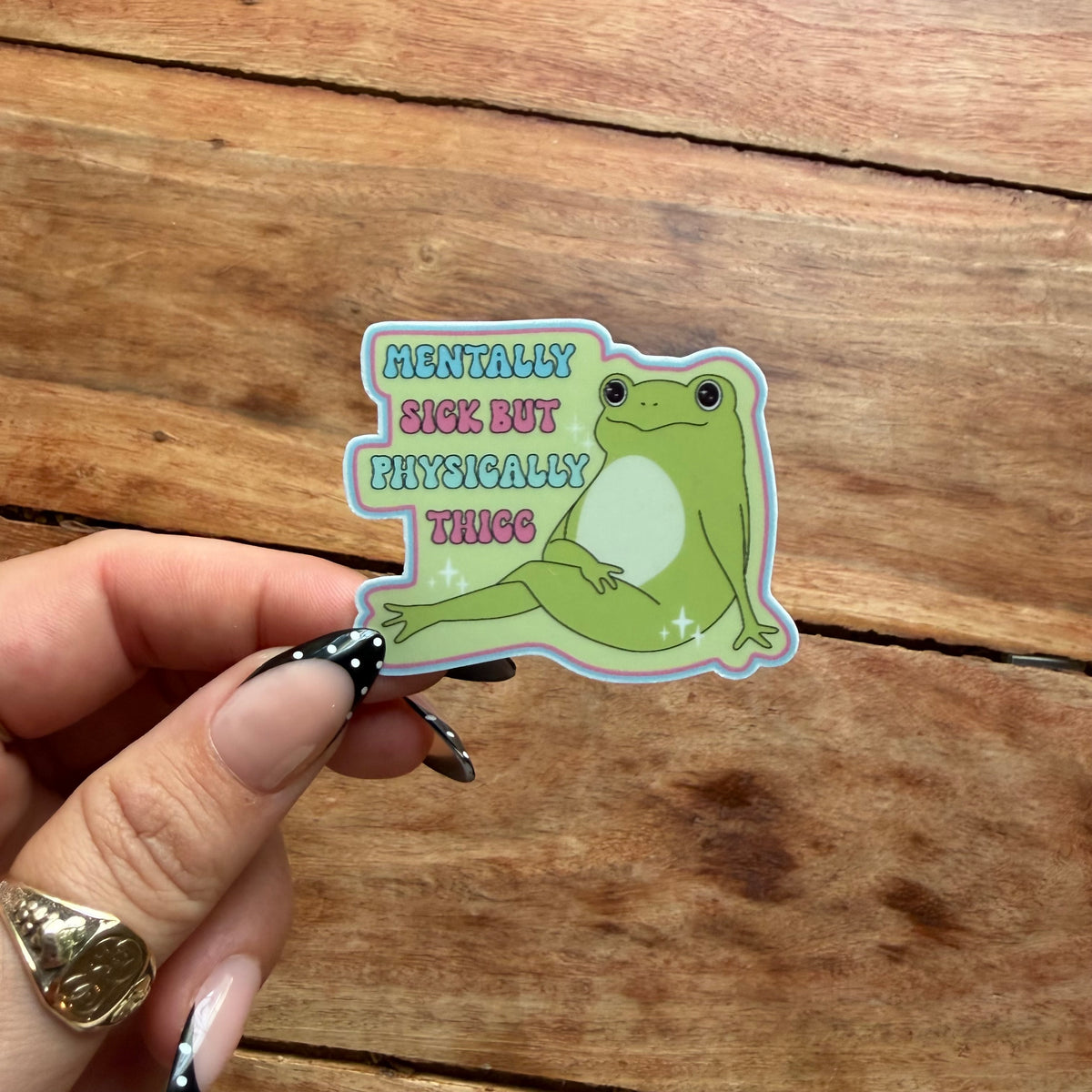 A hand with manicured nails holds a die-cut sticker featuring a green cartoon frog sitting next to the text 'MENTALLY SICK BUT PHYSICALLY THICK' in pastel colors, set against a rustic wooden background.