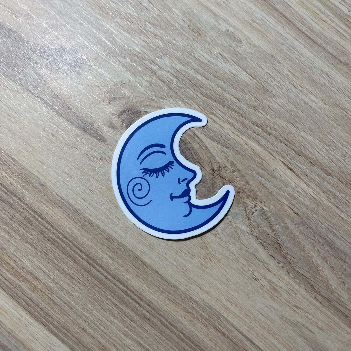 Blue moon-shaped sticker with a face design on a wooden surface