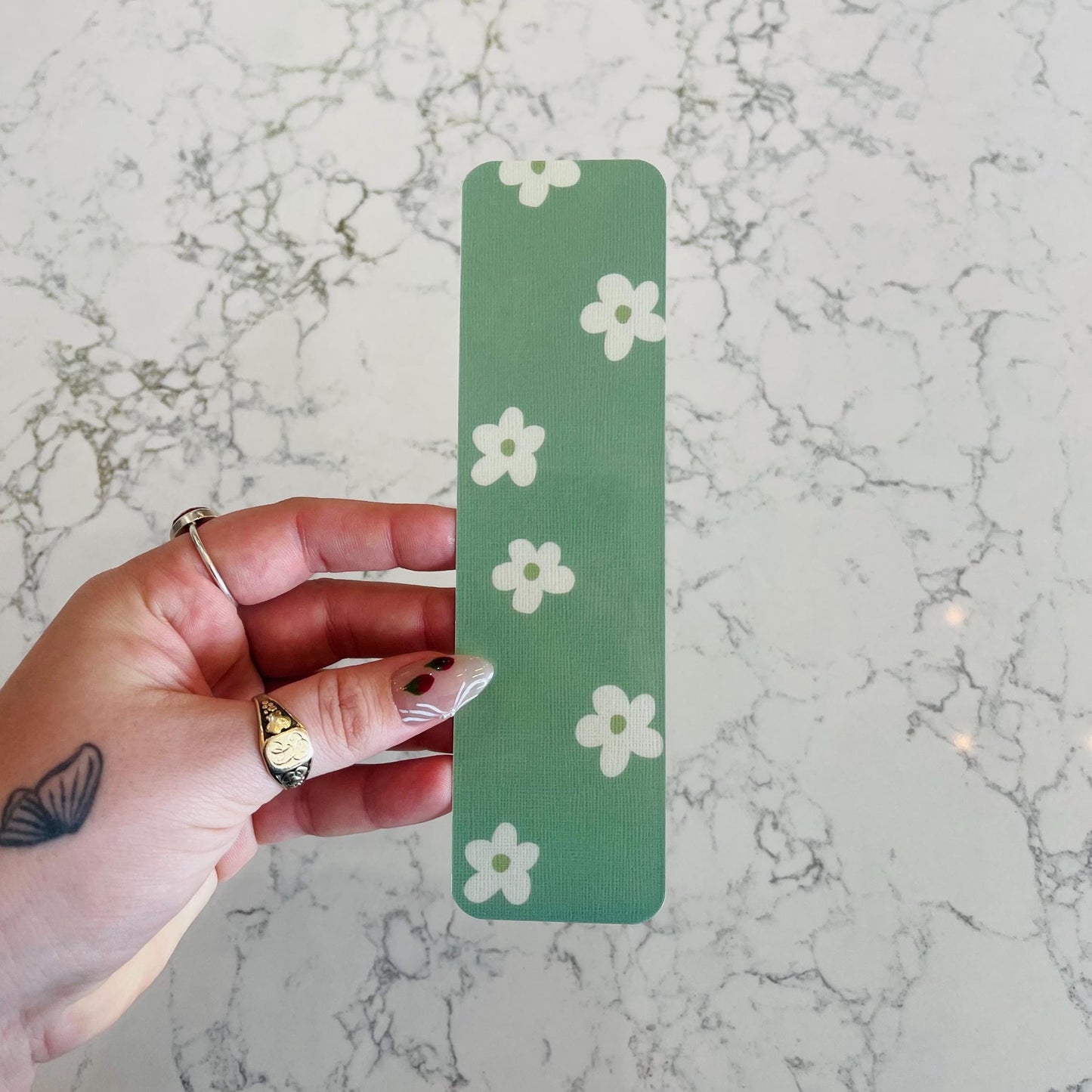 Hand holding a green bookmark with white floral patterns on a marble surface