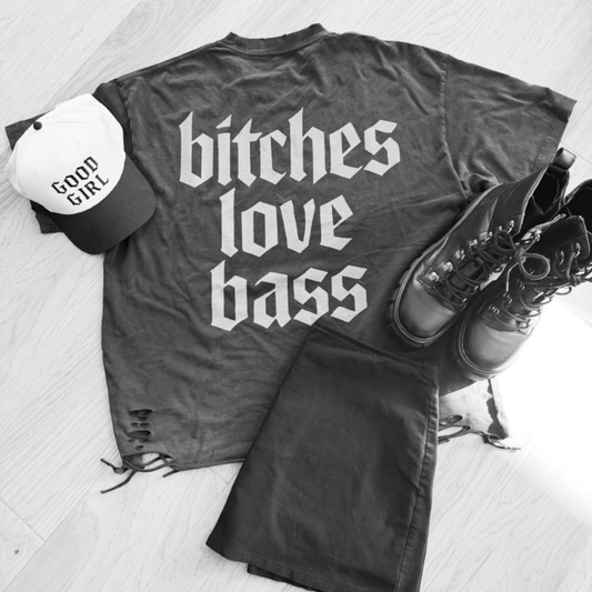 Gray t-shirt with 'bitches love bass' text, white cap, and boots on a light wooden floor.