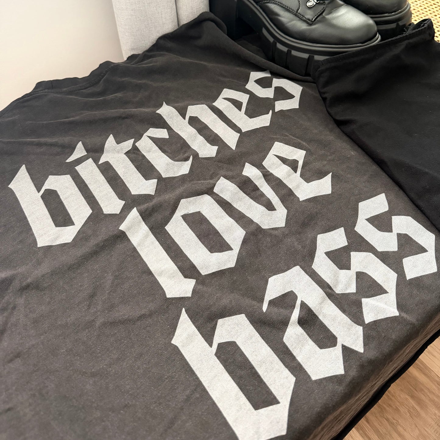 Gray t-shirt with white text 'bitches love bass' and black boots on a light-colored surface.