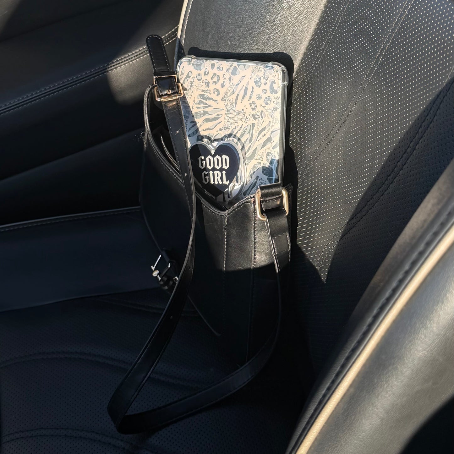 Phone holder with 'good Girl' design attached to a kindle inside a black purse in the seat of a car 