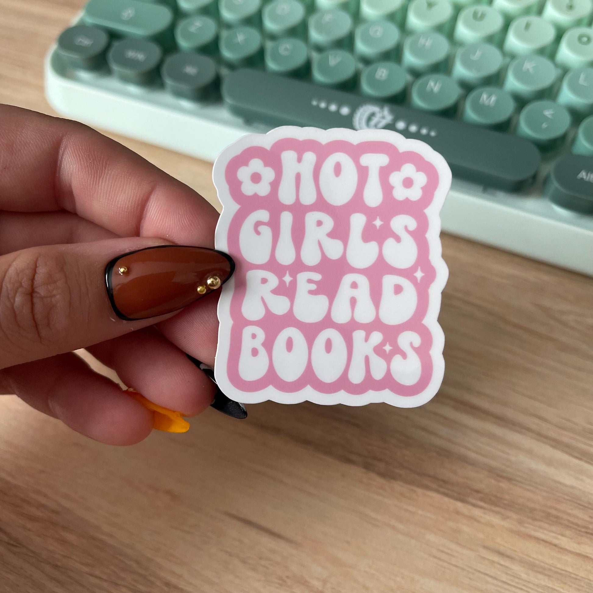 Hand holding a pink sticker with 'Hot Girls Read Books' text in front of a green keyboard.