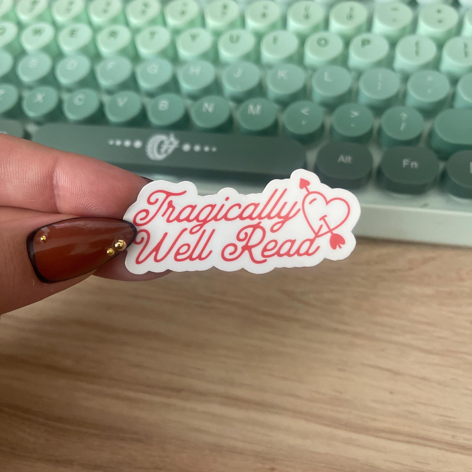Hand holding a 'Tragically Well Read' sticker in front of a green keyboard.