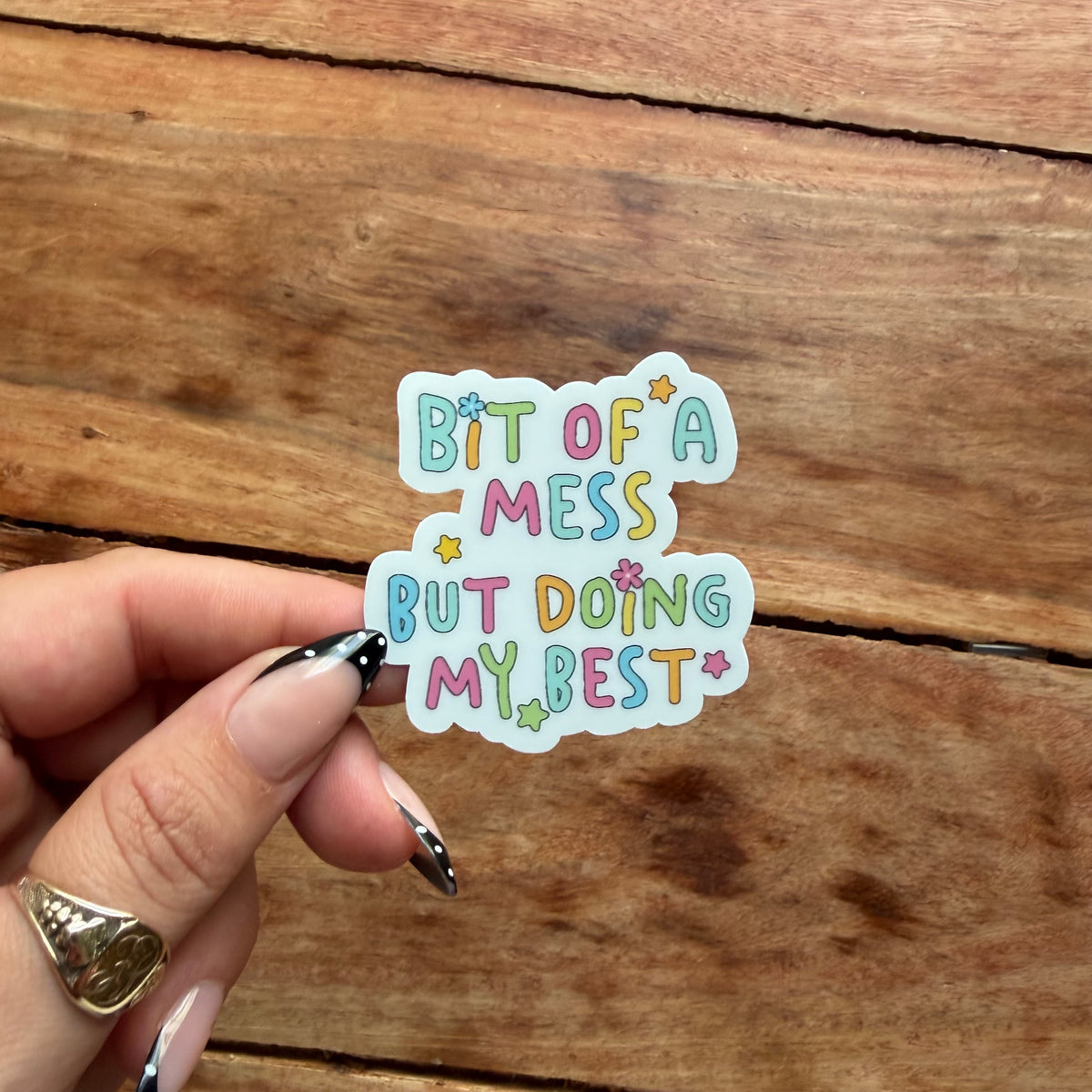 A hand with pink and black nail art holds a die-cut sticker that reads 'BIT OF A MESS BUT DOING MY BEST' in pastel multicolor letters on a white background, set against a rustic wooden surface.