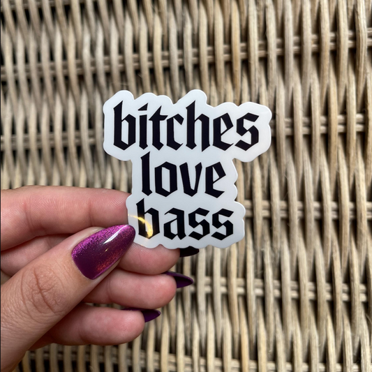 Sticker with 'bitches love bass' text held by a hand against a wicker background