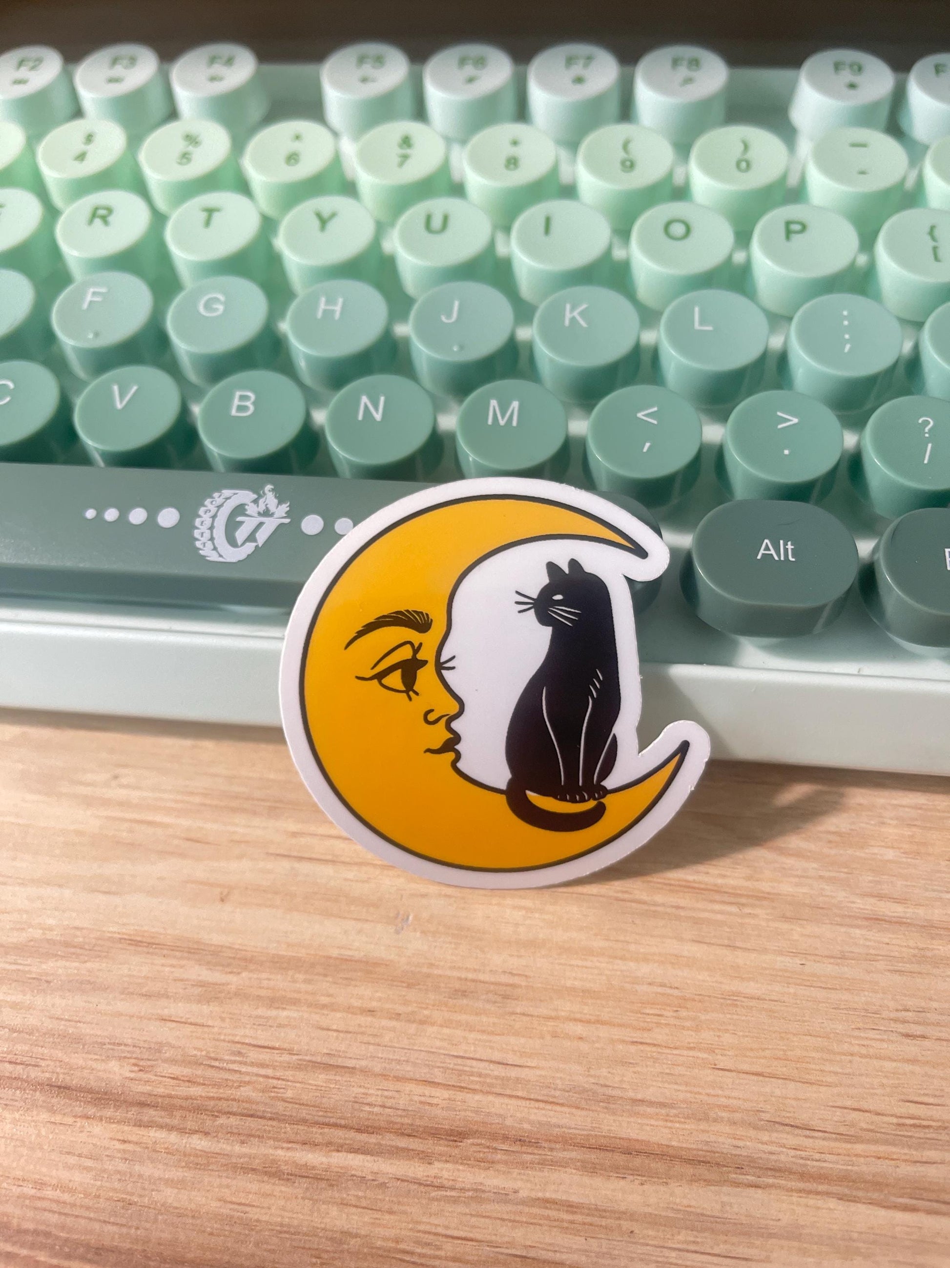 Sticker with a crescent moon face design on a wooden surface in front of a green keyboard.