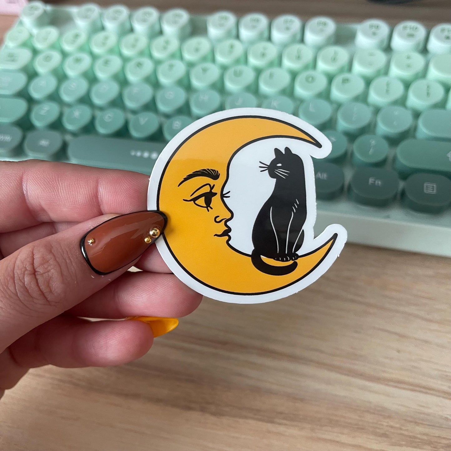 Hand holding a sticker with a cat on a moon design in front of a keyboard.