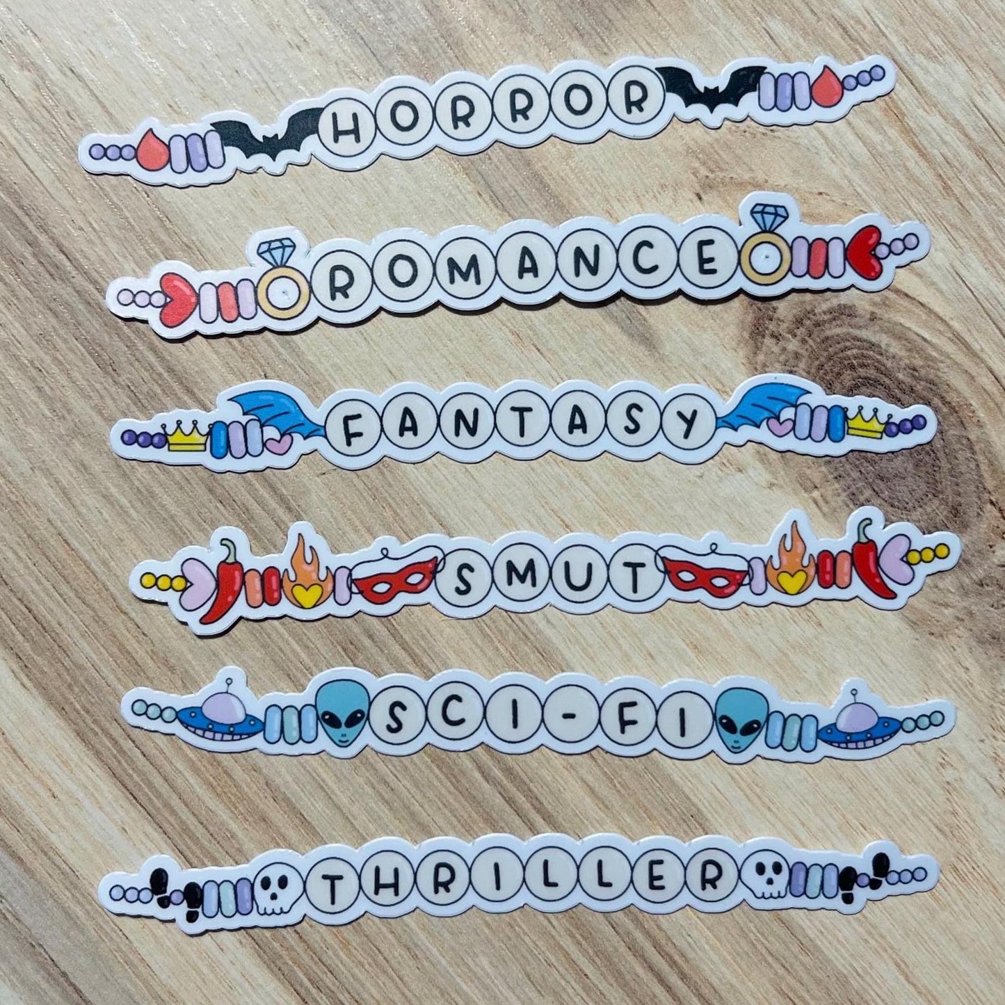 Stickers labeled 'Horror', 'Romance', 'Fantasy', 'Smut', 'Sci-Fi', and 'Thriller' on a wooden surface.