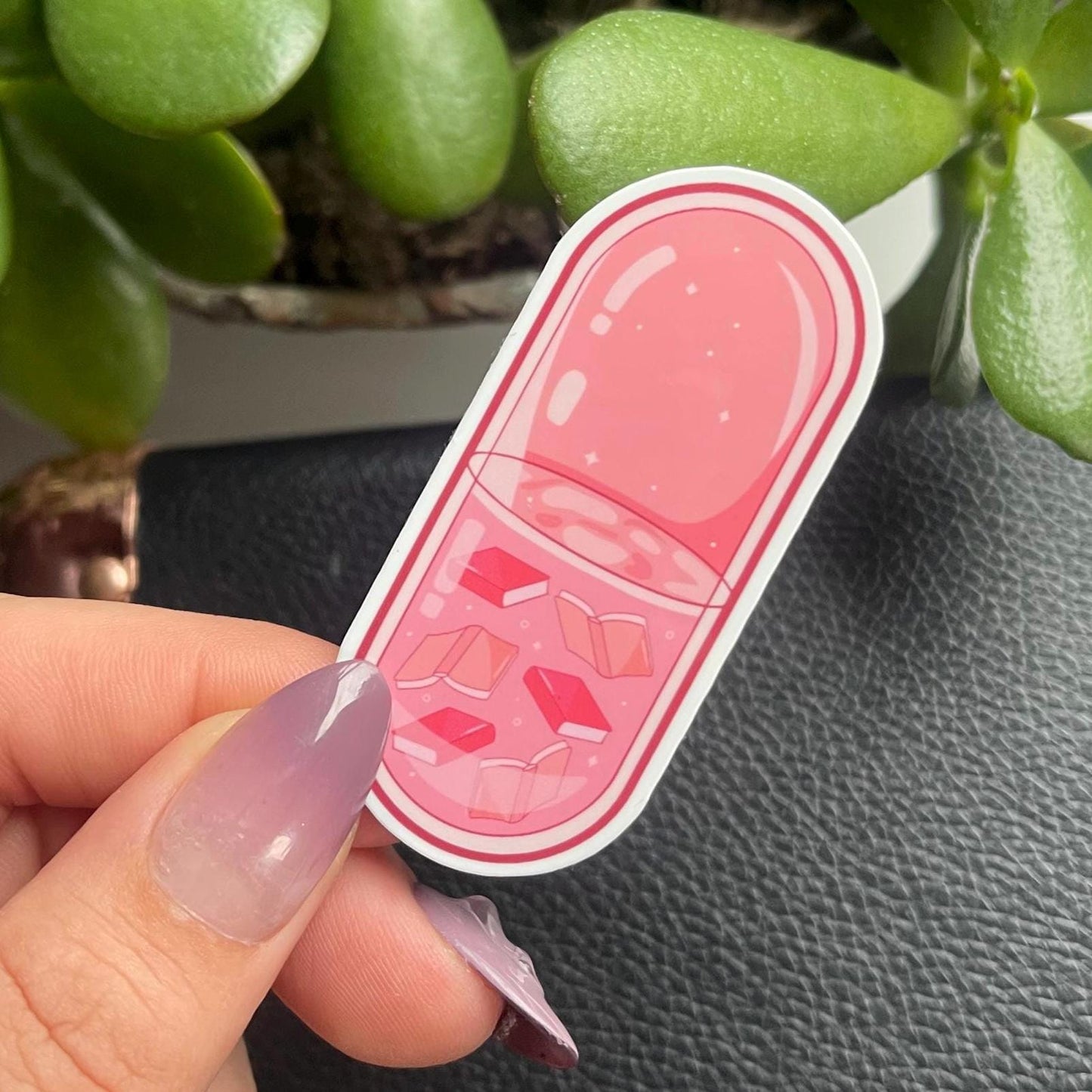 Pink pill-shaped sticker held by a hand with a plant in the background