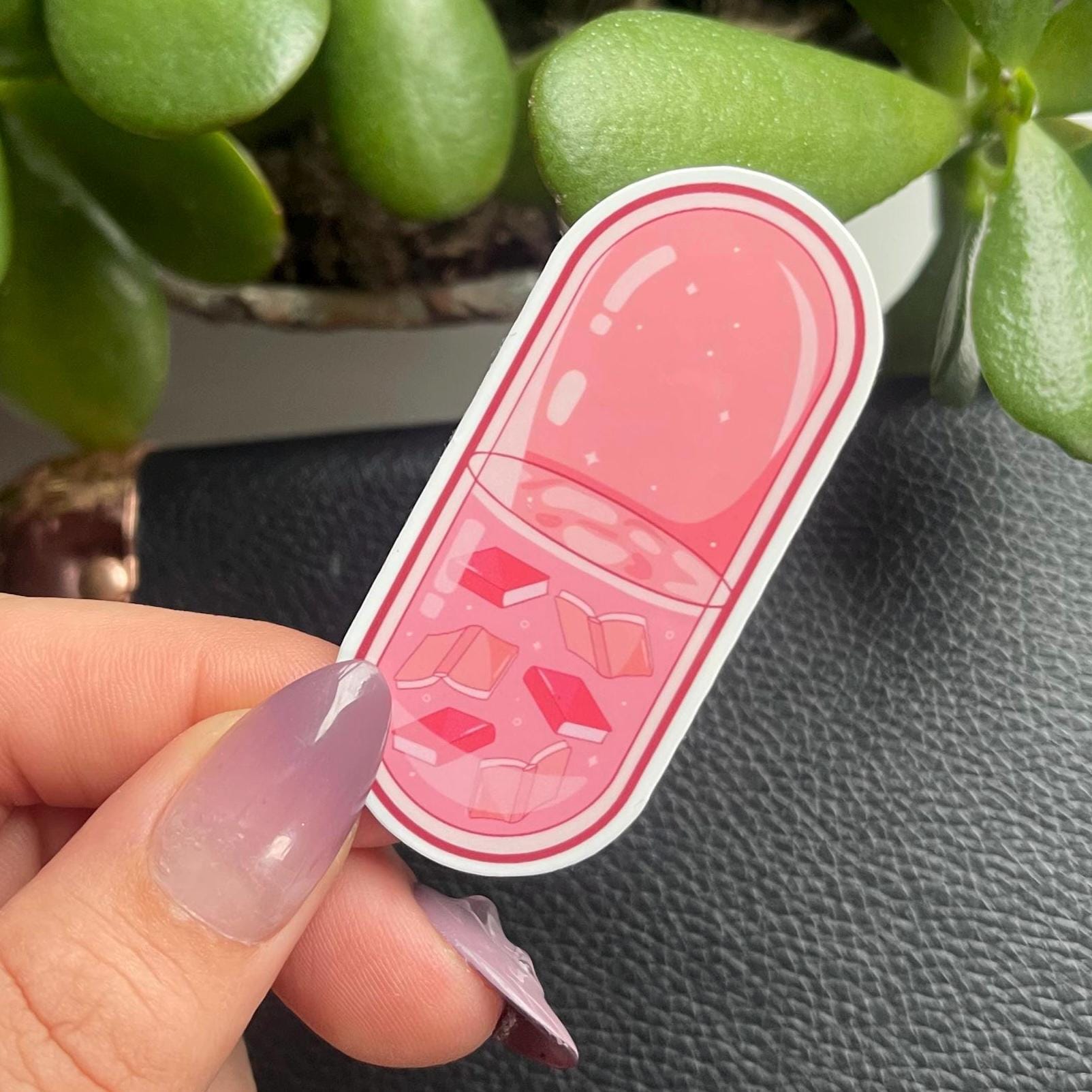 Pink pill-shaped sticker held by a hand with a plant in the background