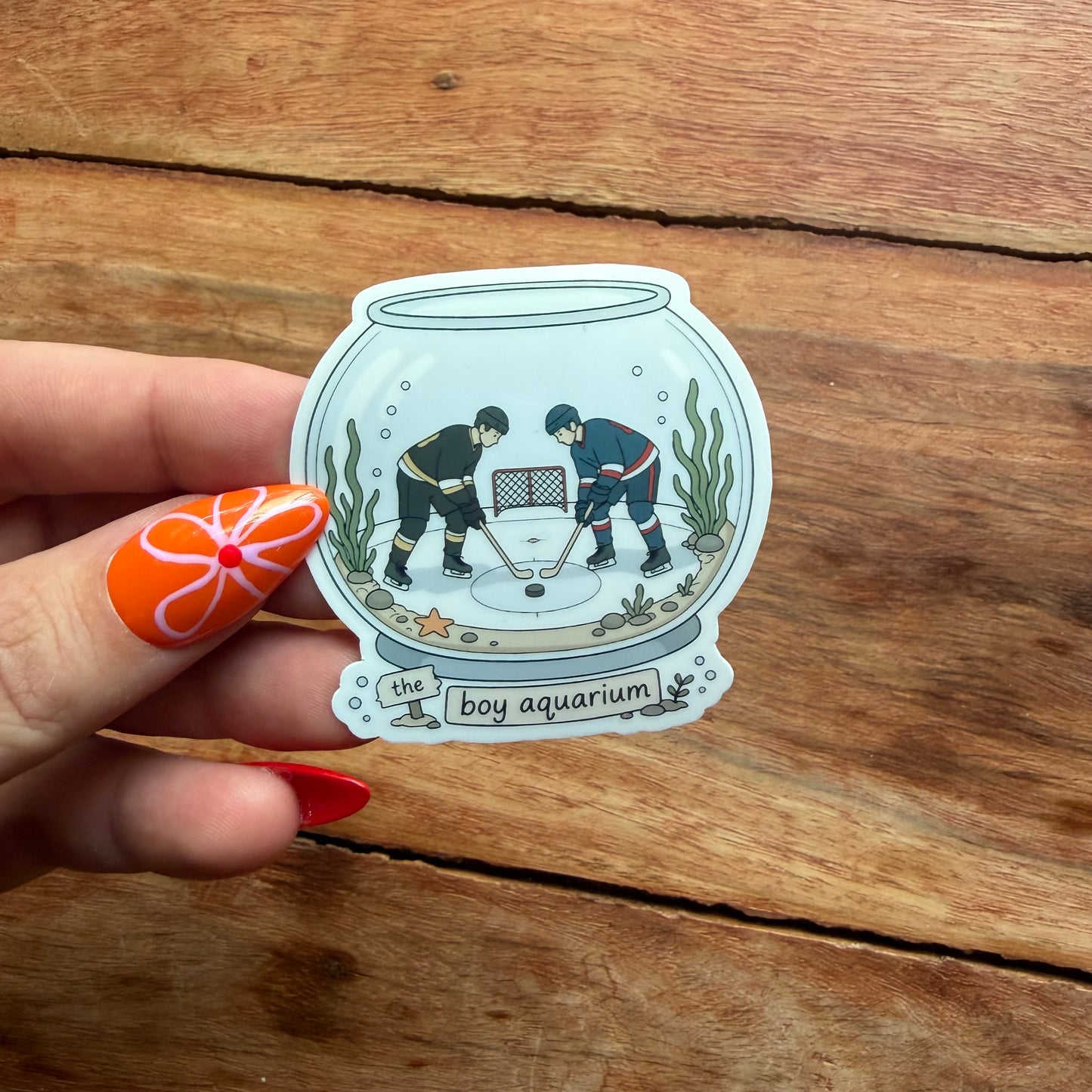 Sticker of two people playing hockey inside an aquarium held by a hand on a wooden surface