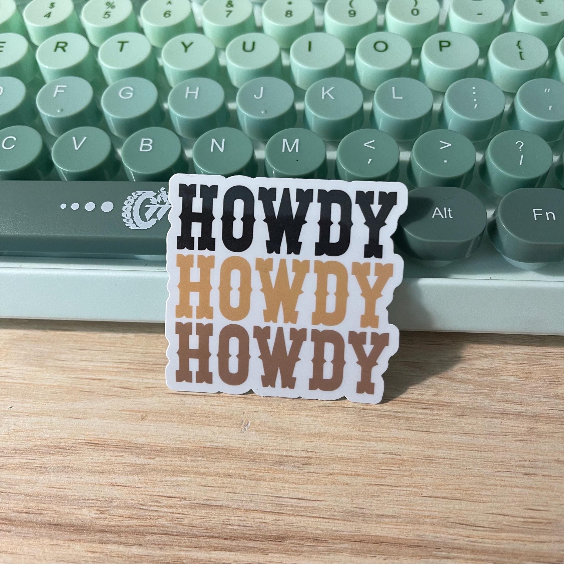 Sticker with 'HOWDY' text on a wooden surface with a typewriter in the background