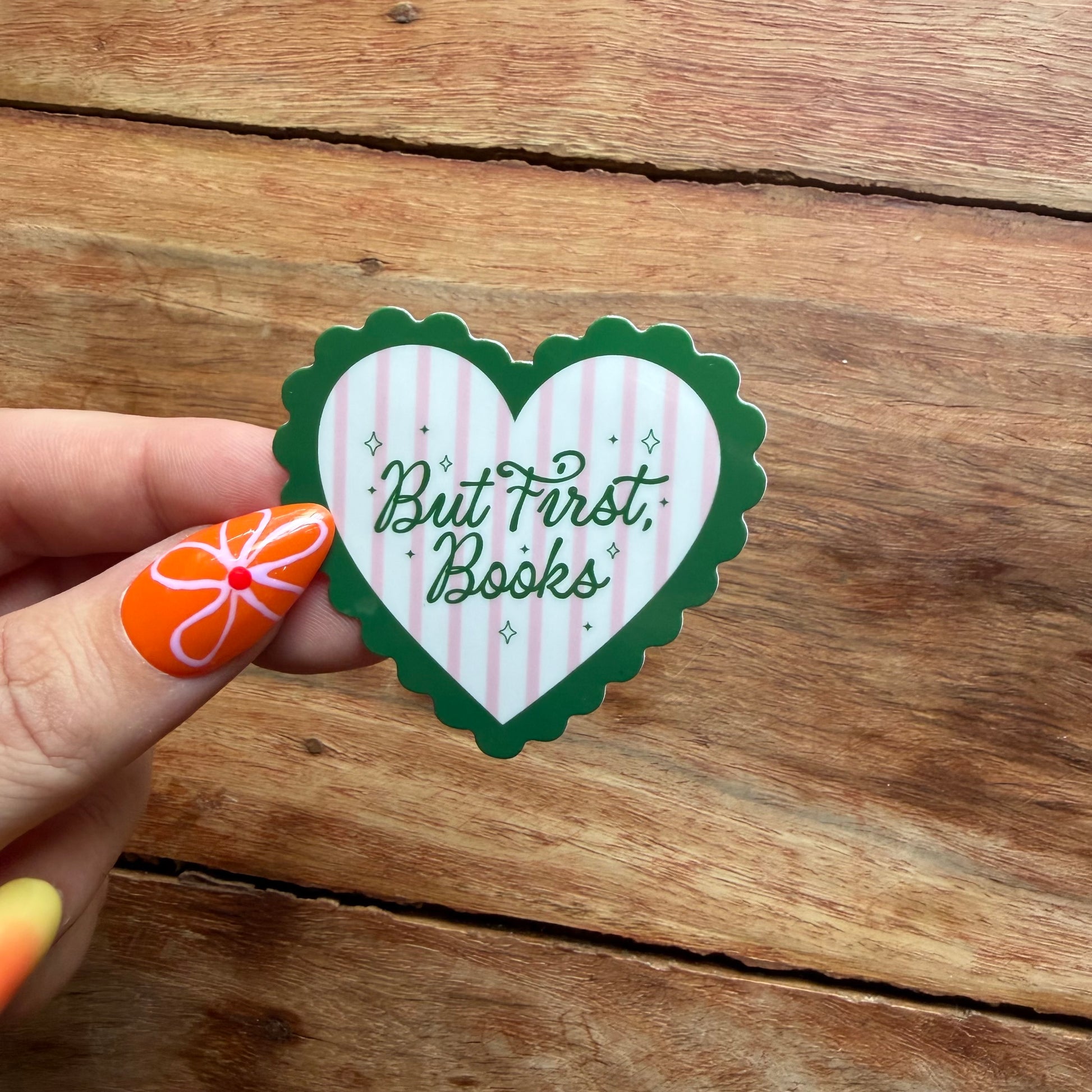 Heart-shaped sticker with 'But First, Books' text held by a hand on a wooden surface