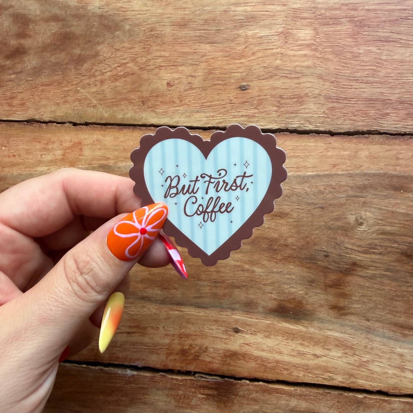 Heart-shaped sticker with 'But First, Coffee' text held by a hand on a wooden surface