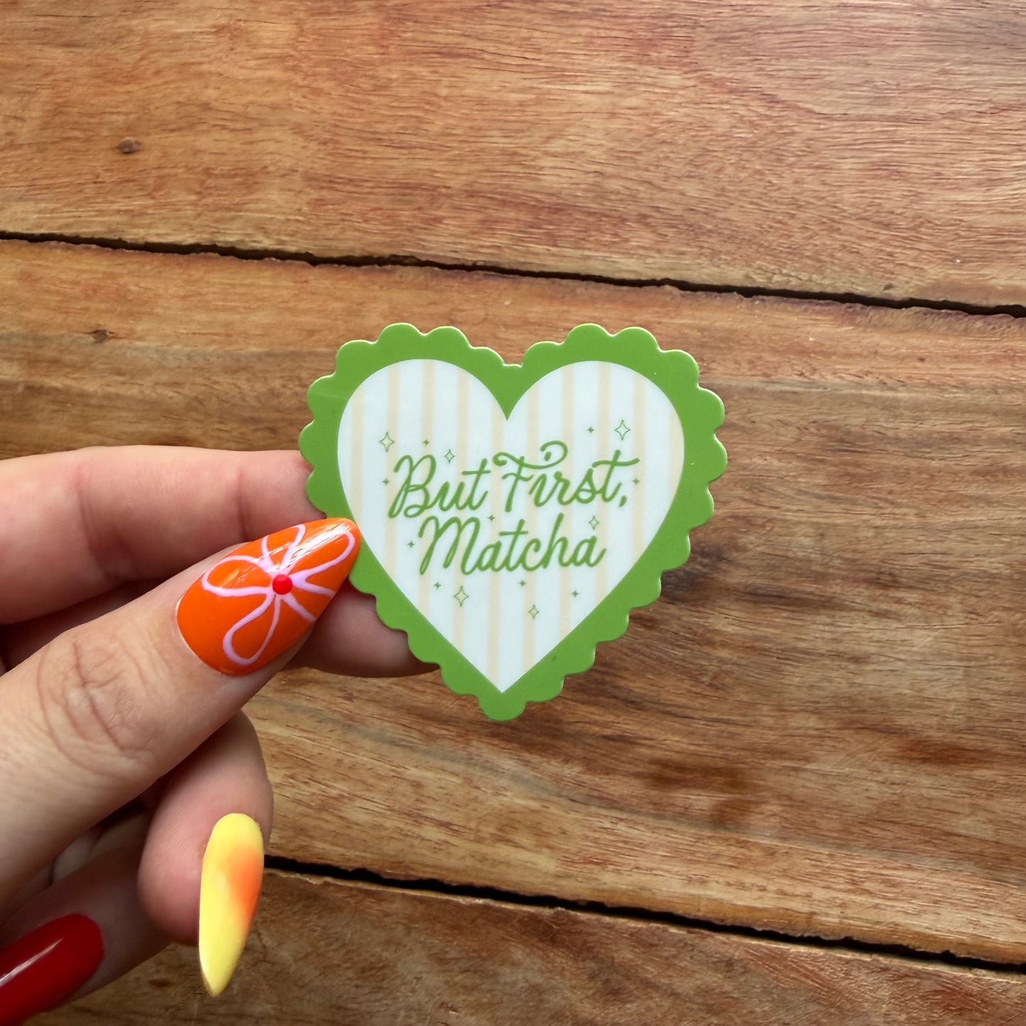 Heart-shaped sticker with 'But First, Matcha' text held by a hand on a wooden surface