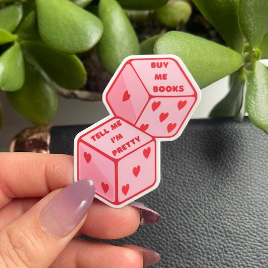 Pink dice-shaped sticker with text held by a hand in front of green plants.