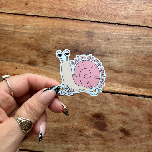 A hand with black and white manicured nails holds a die-cut sticker featuring a pink snail with the words 'Calm down weirdo' around it, set against a rustic wooden background.