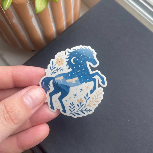 Hand holding a blue horse sticker with floral and celestial design against a dark background.