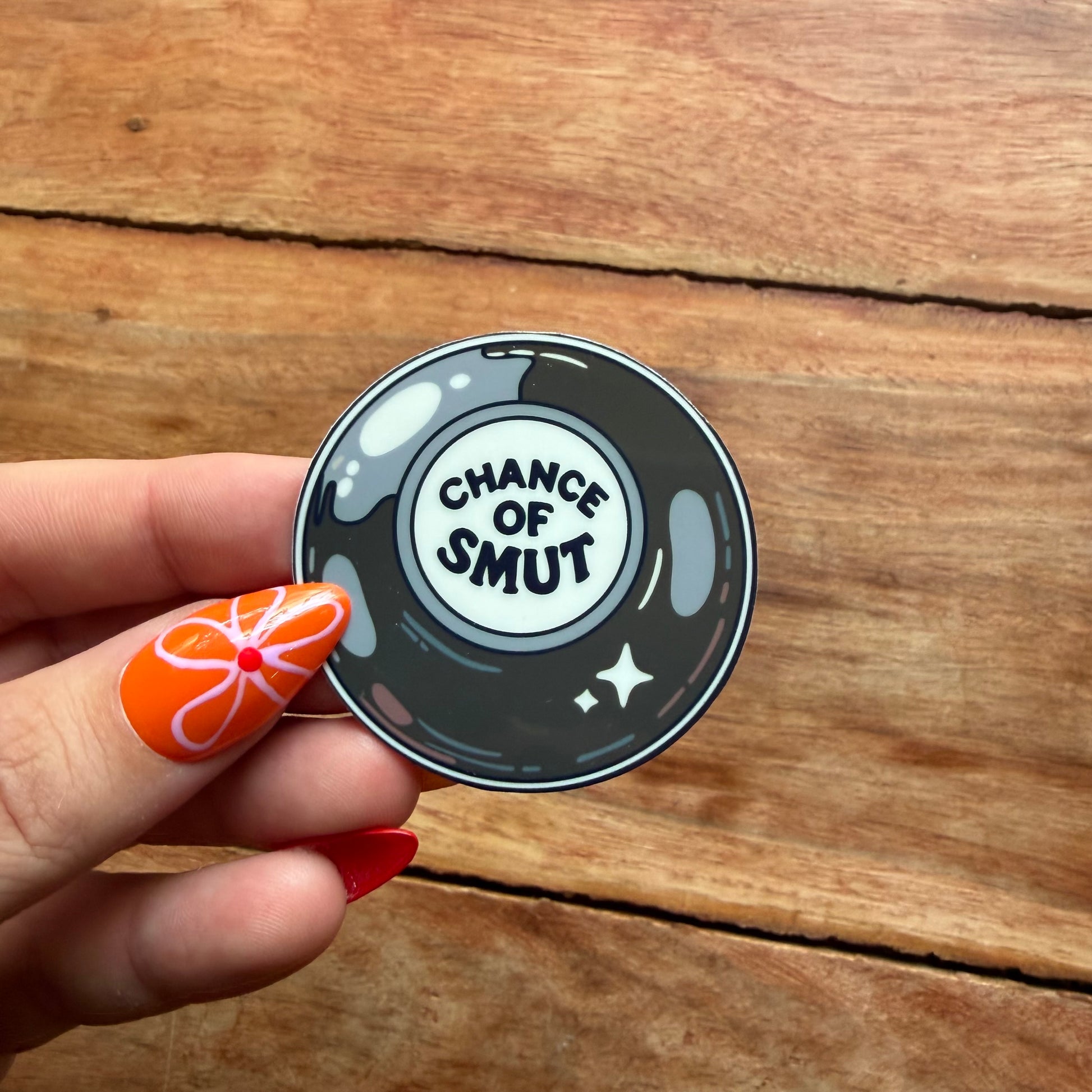 Sticker with 'Chance of Smut' text held by a hand on a wooden surface