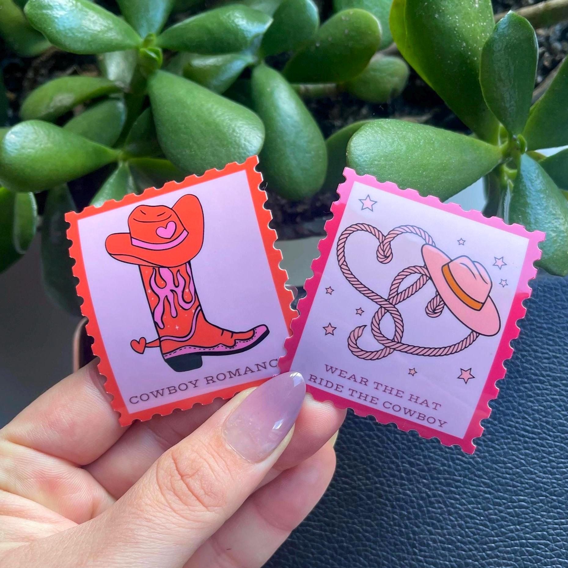 Two colorful stickers with cowboy-themed designs held by a hand against a green plant background.