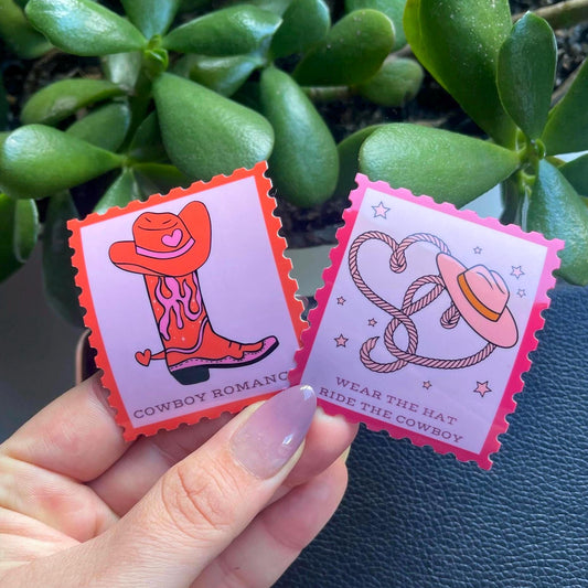 Two colorful stickers with cowboy-themed designs held by a hand against a green plant background.