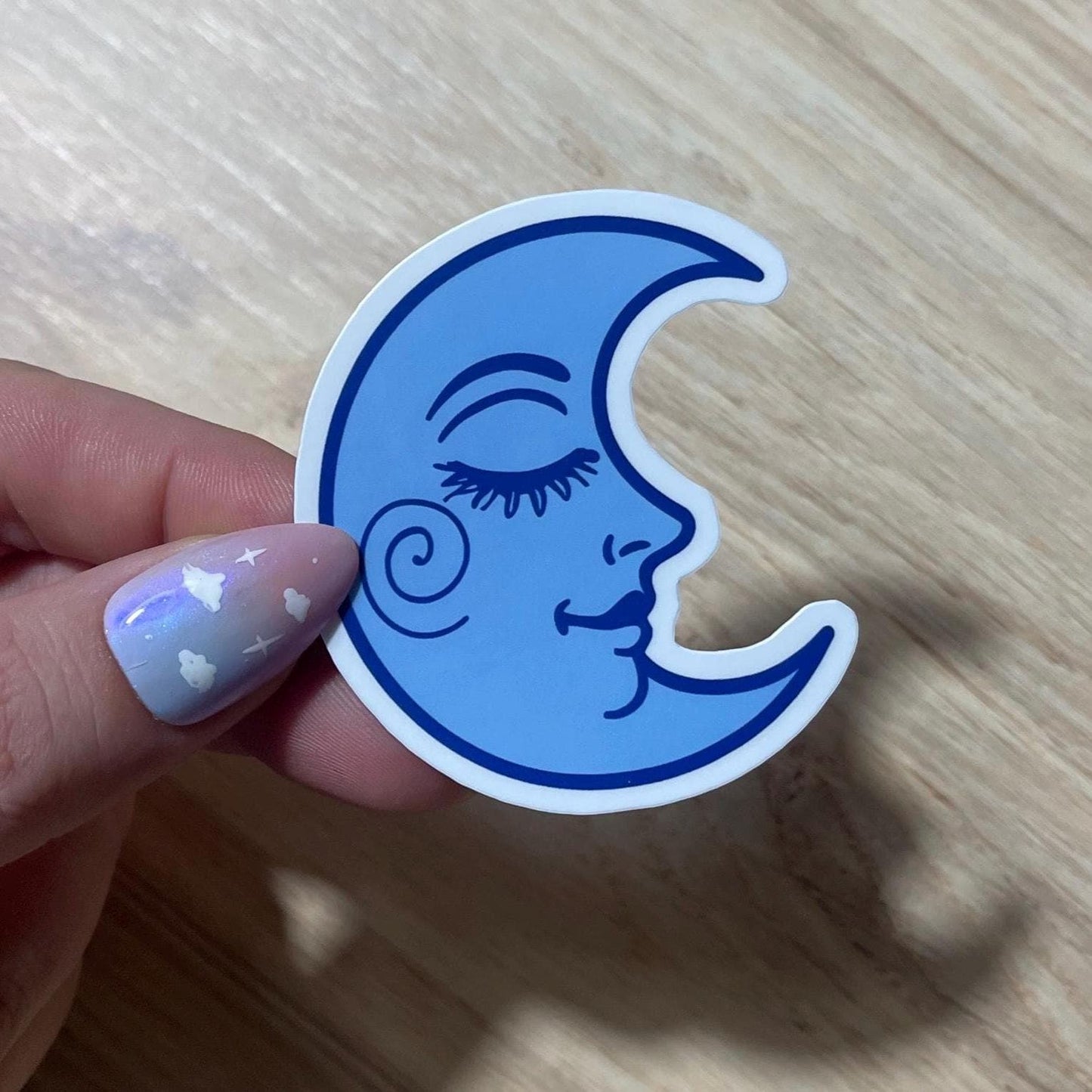 Blue crescent moon sticker with a face held by a hand on a wooden surface