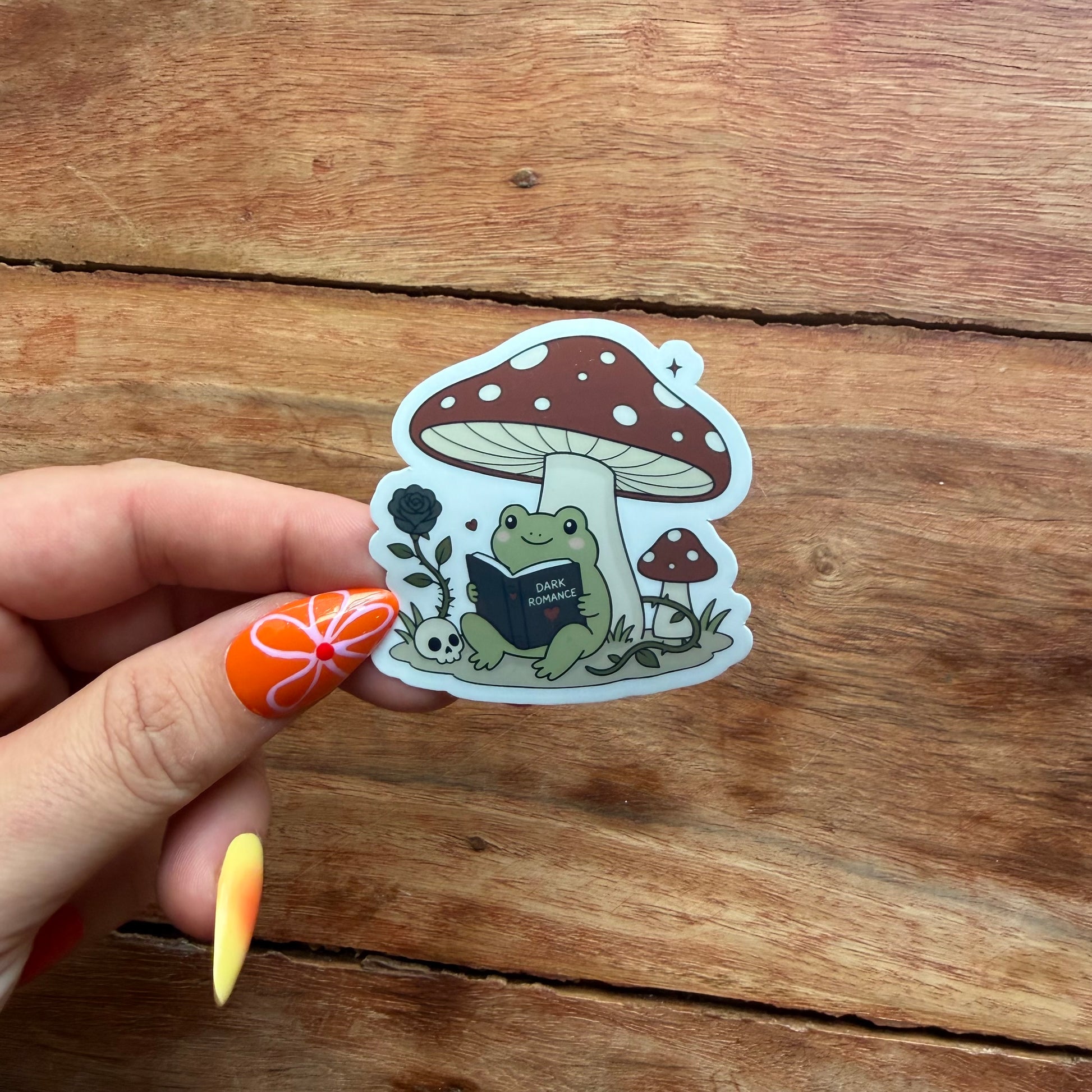 Sticker of a frog reading a book under a mushroom held by a hand on a wooden surface