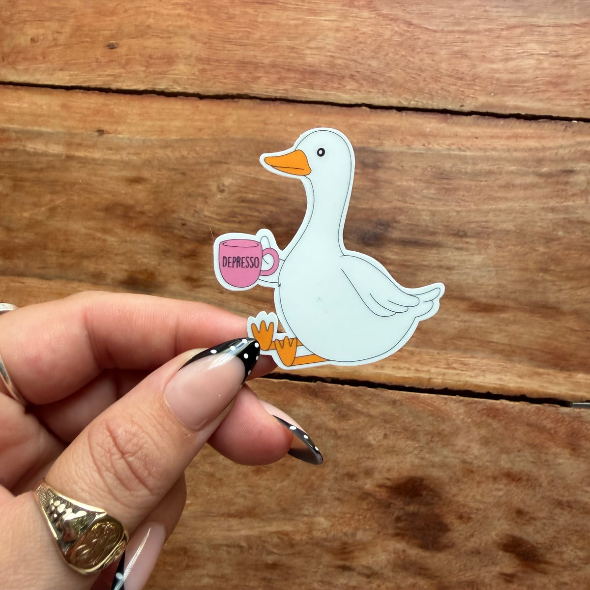 A hand with manicured nails holds a die-cut sticker of a white goose holding a pink espresso mug, set against a rustic wooden background.