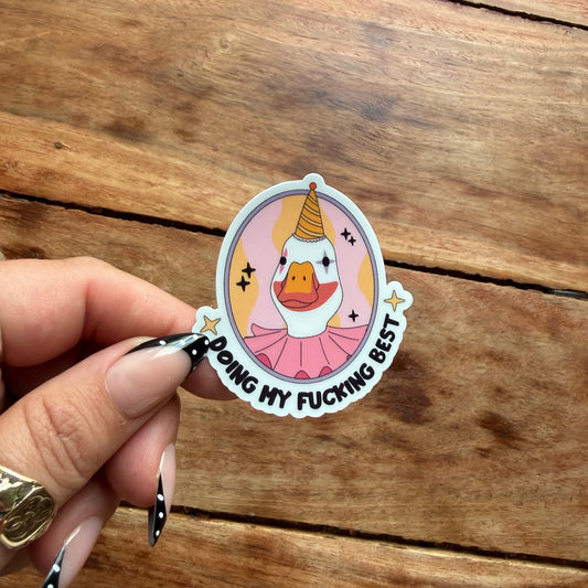 A hand with black and white patterned nail polish holds a die-cut sticker featuring a cartoon duck in a pink tutu and party hat with the text 'DOING MY FUCKING BEST' on a rustic wooden background.