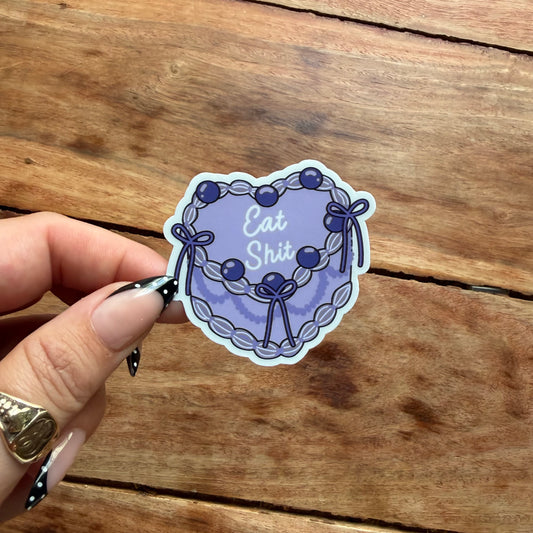 A hand with manicured nails holds a die-cut sticker featuring a lavender and purple floral wreath with the text 'Eat Shit' in white script, set against a rustic wooden background.