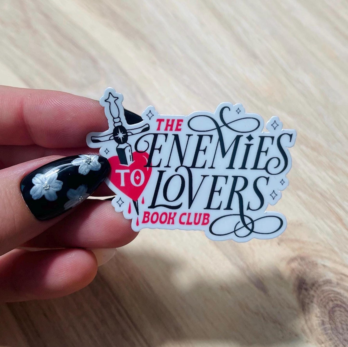 Sticker for 'The Enemies to Lovers Book Club' held by a hand with black nail polish on a wooden surface.