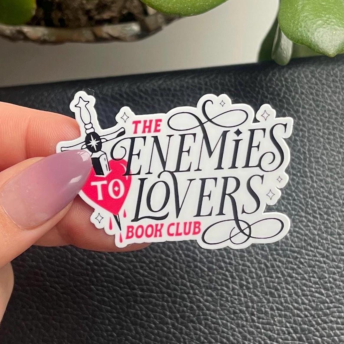 Sticker with 'The Enemies to Lovers Book Club' text held by a hand on a black surface.