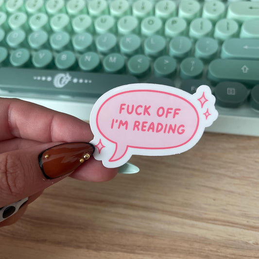 Hand holding a sticker with 'FUCK OFF I'M READING' text in front of a keyboard.