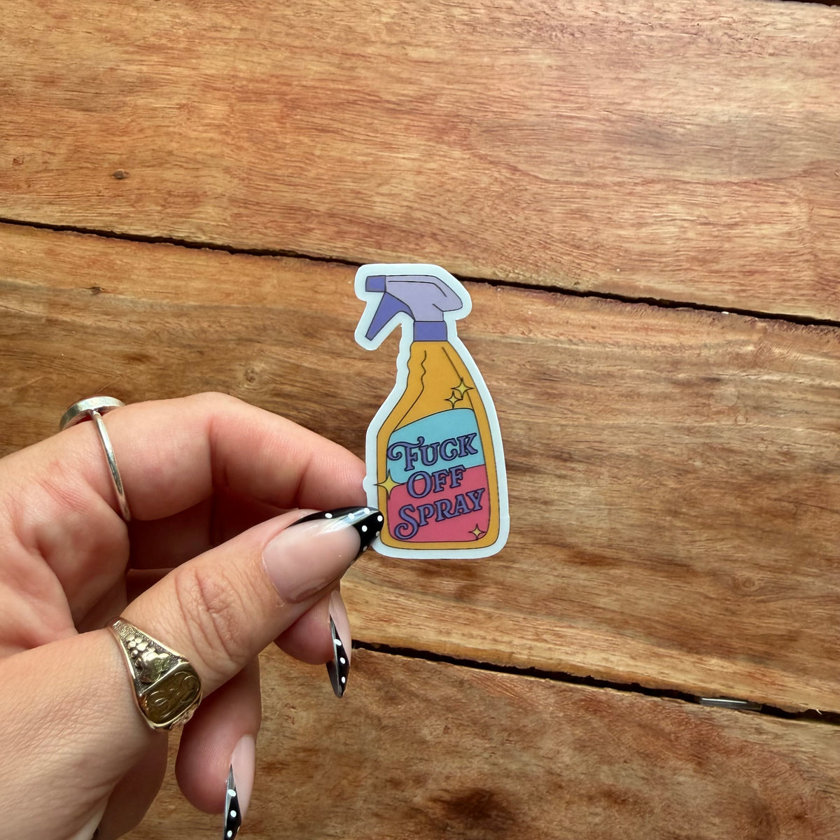 A hand with silver rings holds a die-cut sticker shaped like a spray bottle labeled 'FUCK OFF SPRAY' against a rustic wooden background.