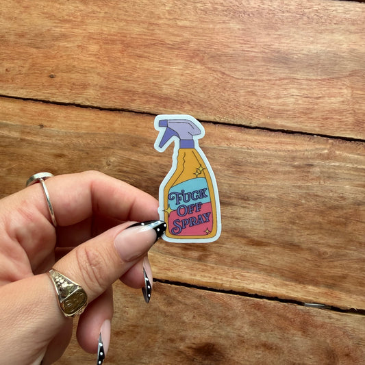 A hand with silver rings holds a die-cut sticker shaped like a spray bottle labeled 'FUCK OFF SPRAY' against a rustic wooden background.