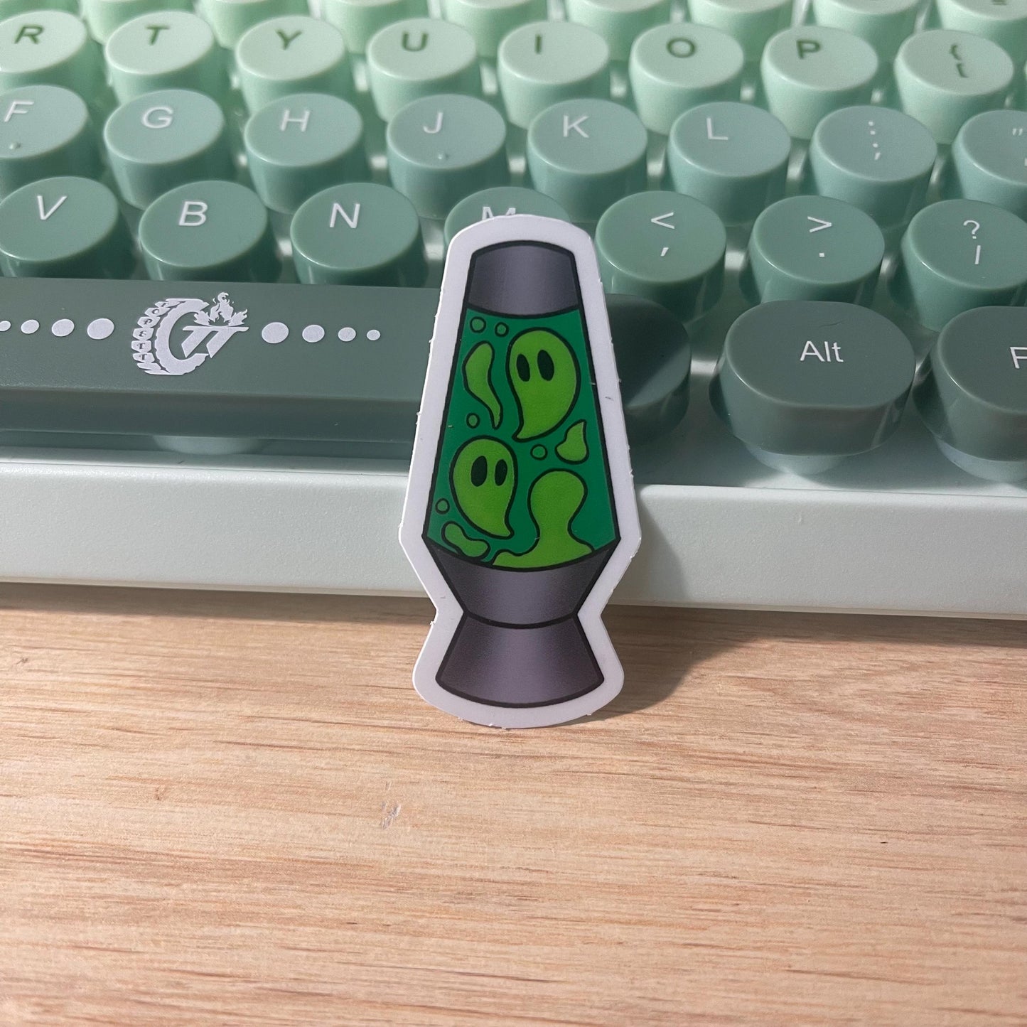 Sticker of a green lava lamp with ghost design on a wooden surface in front of a keyboard.