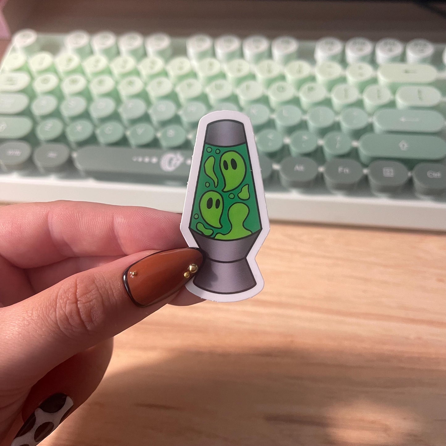 Hand holding a sticker of a green lava lamp with ghost design in front of a keyboard.