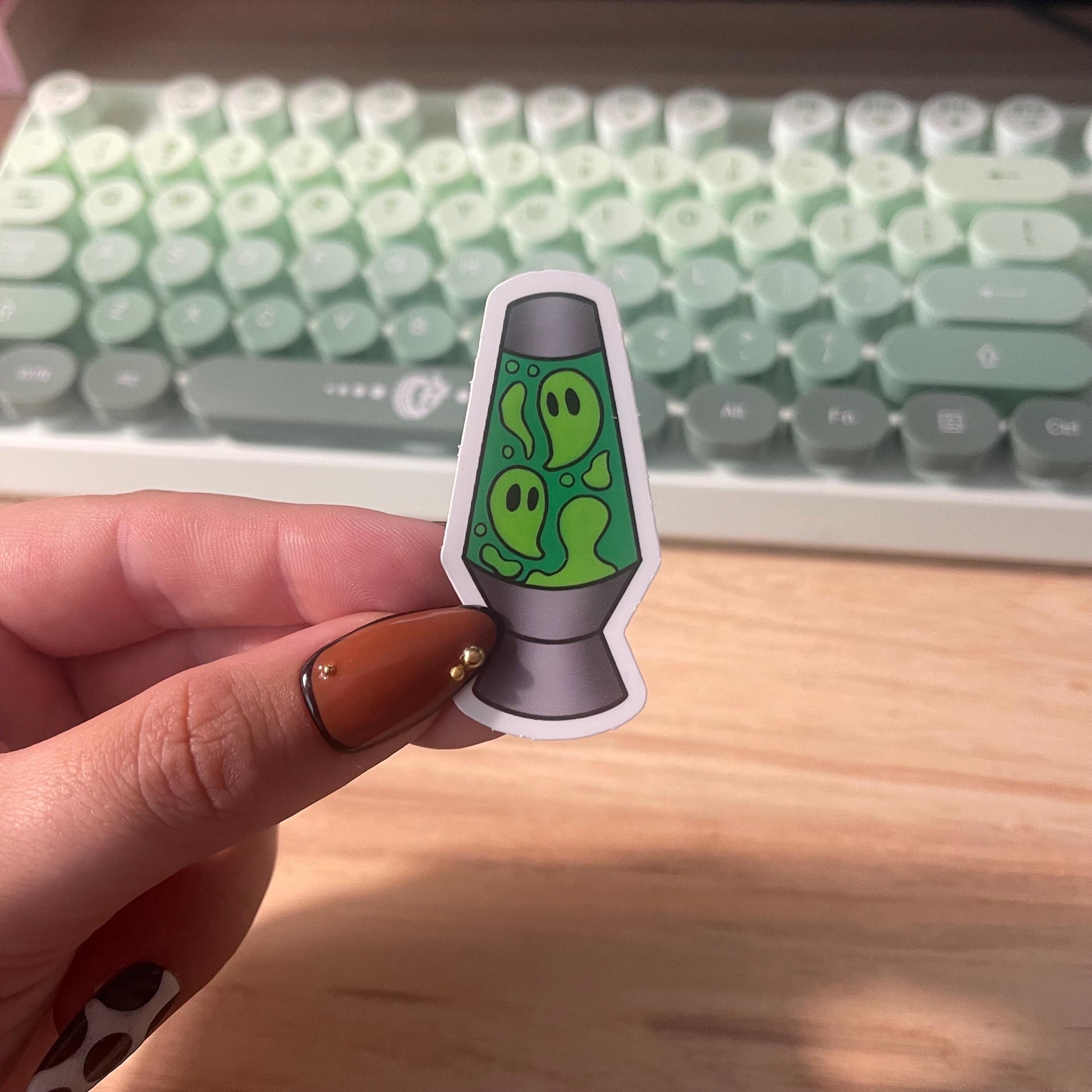 Hand holding a sticker of a green lava lamp with ghost design in front of a keyboard.