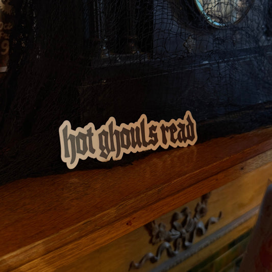 A close-up of a black textured journal or notebook cover with a small die-cut bookmark that reads 'hot ghouls read' in a stylized font, next to a wooden surface.