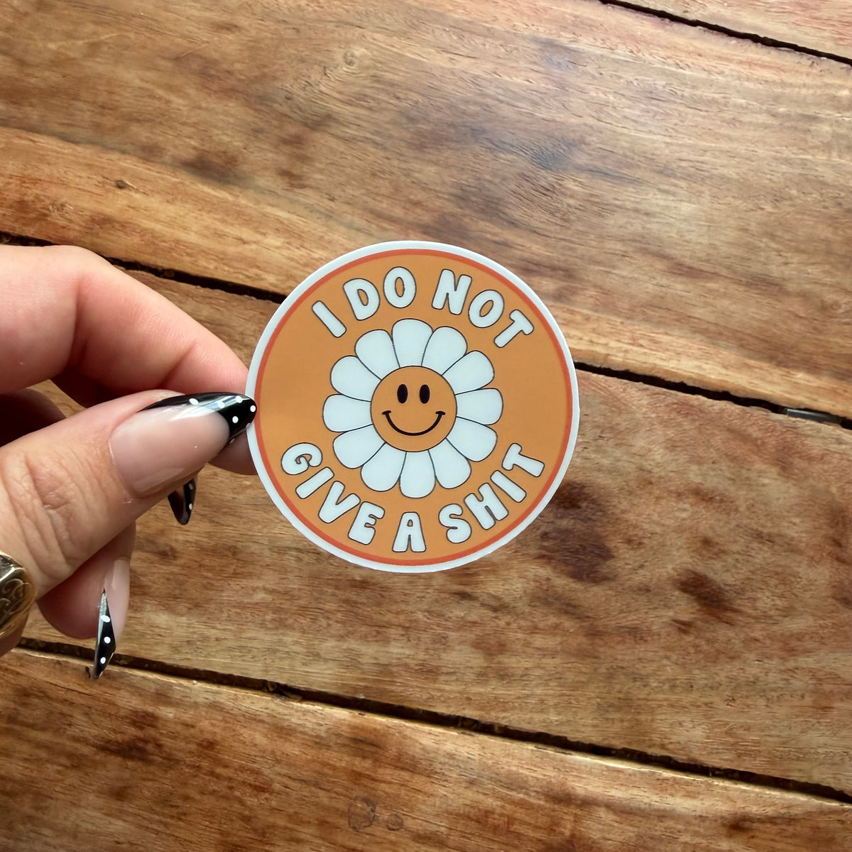 A hand with black and white nail art holds a round sticker featuring a smiling daisy on an orange background with the text 'I DO NOT GIVE A SHIT'. The sticker is photographed against a rustic wooden plank surface.