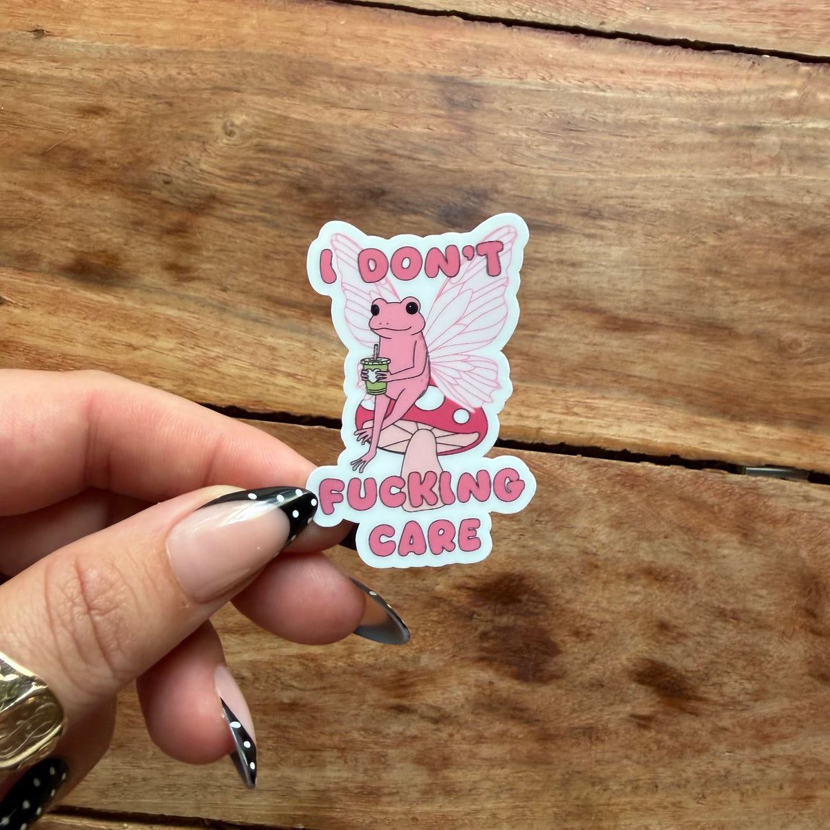 A hand with manicured nails holds a die-cut sticker featuring a pink fairy holding a bouquet next to a red-and-white mushroom, with the text 'I DON'T FUCKING CARE' in pink lettering, set against a rustic wooden background.