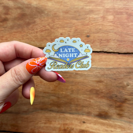 Hand holding a 'Late Night Readers Club' sticker against a wooden background