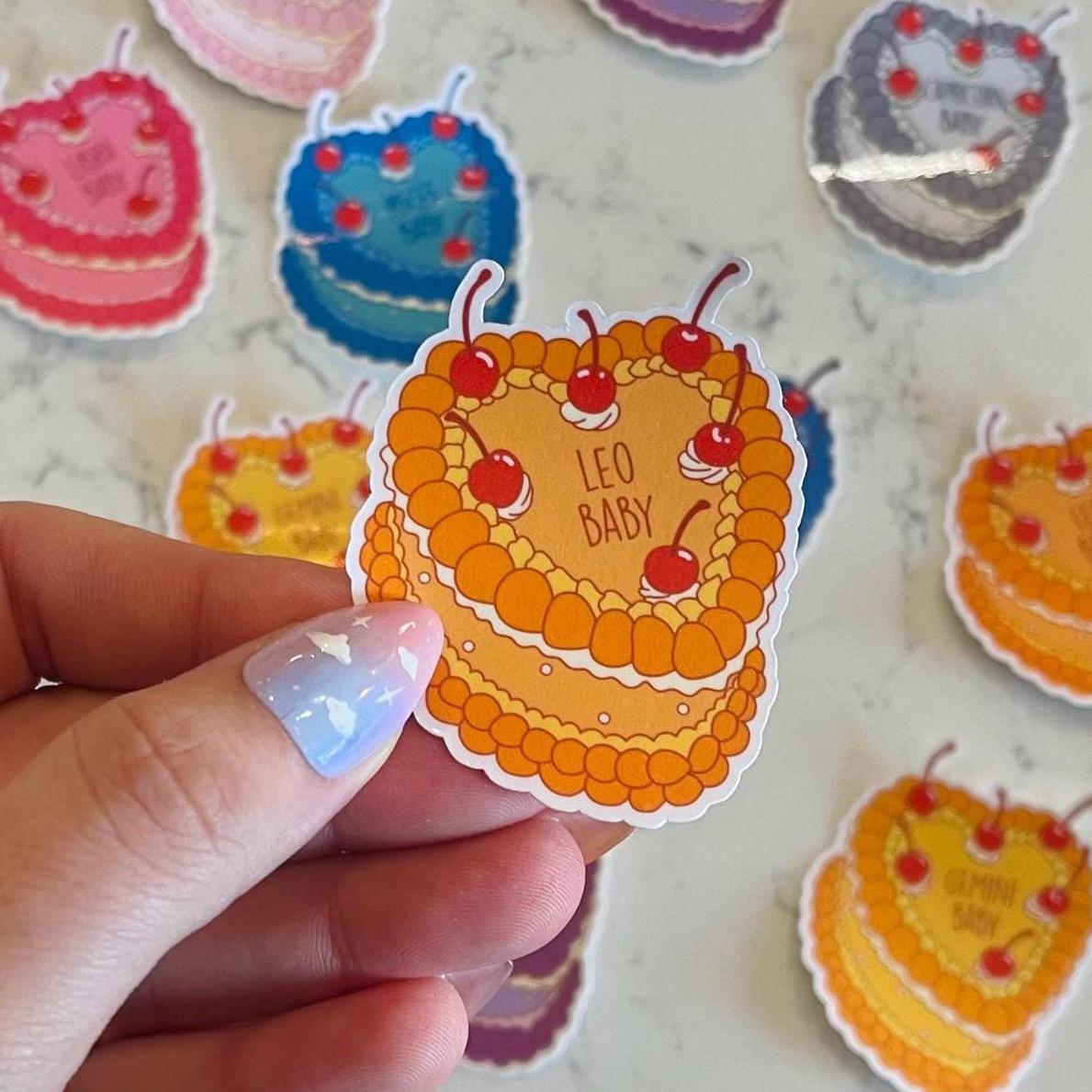 Hand holding a colorful cake-shaped sticker with 'Leo Baby' text, surrounded by similar stickers on a light background.