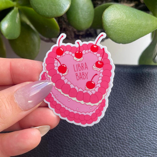 Sticker with 'Libra Baby' text held by a hand against a plant background